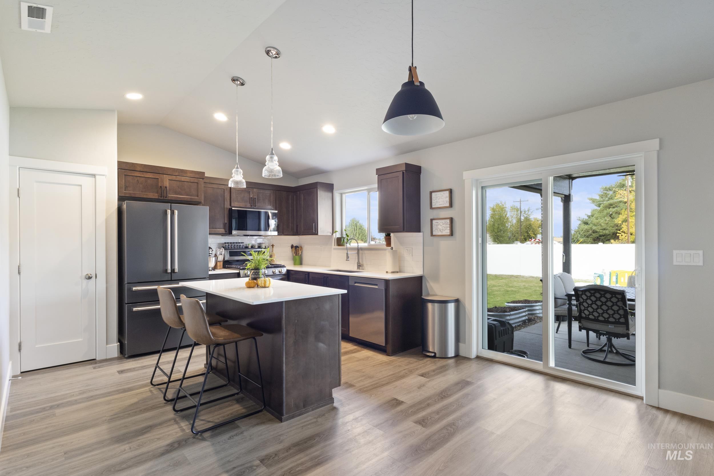 Kitchen featuring dark brown cabinets, vaulted ceiling, appliances with stainless steel finishes, pendant lighting, and a kitchen bar