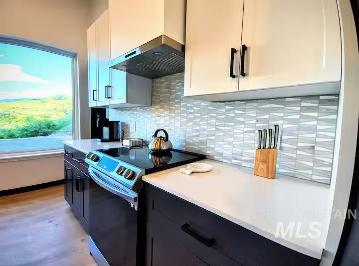 Kitchen featuring stainless steel electric stove, ventilation hood, light countertops, decorative backsplash, and white cabinets