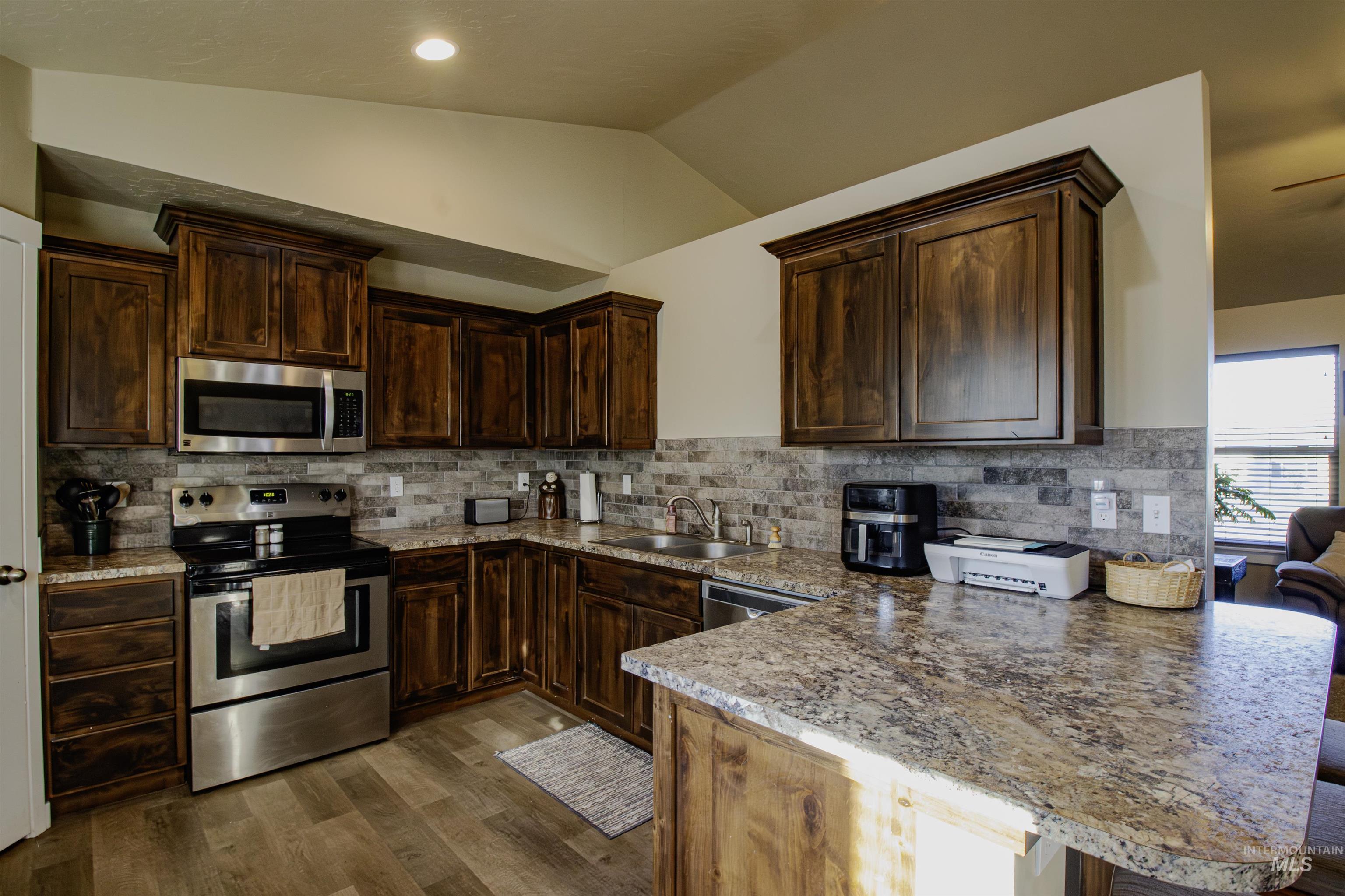 Kitchen featuring a peninsula, stainless steel appliances, dark brown cabinets, vaulted ceiling, and decorative backsplash