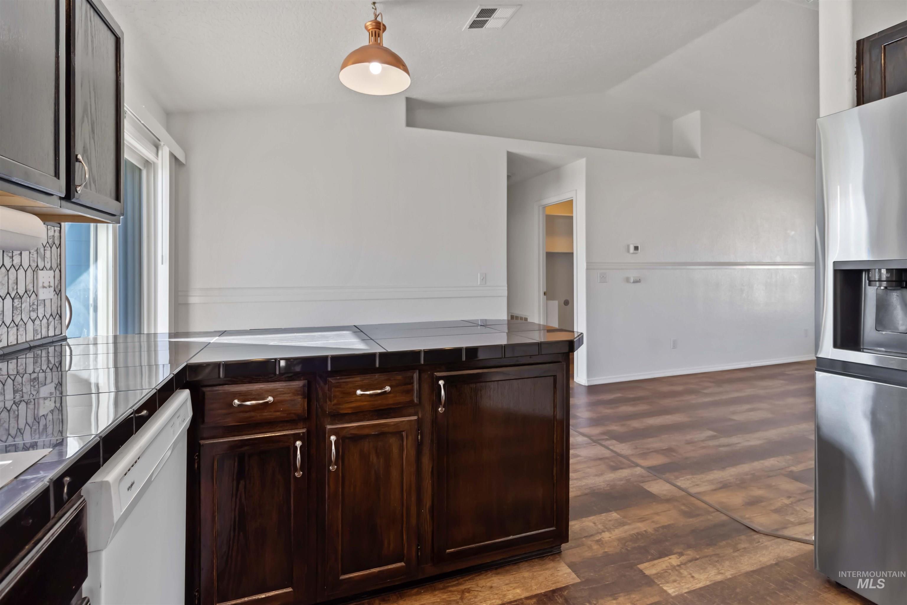 Kitchen with dark brown cabinets, stainless steel fridge with ice dispenser, dark wood-style flooring, white dishwasher, and hanging light fixtures