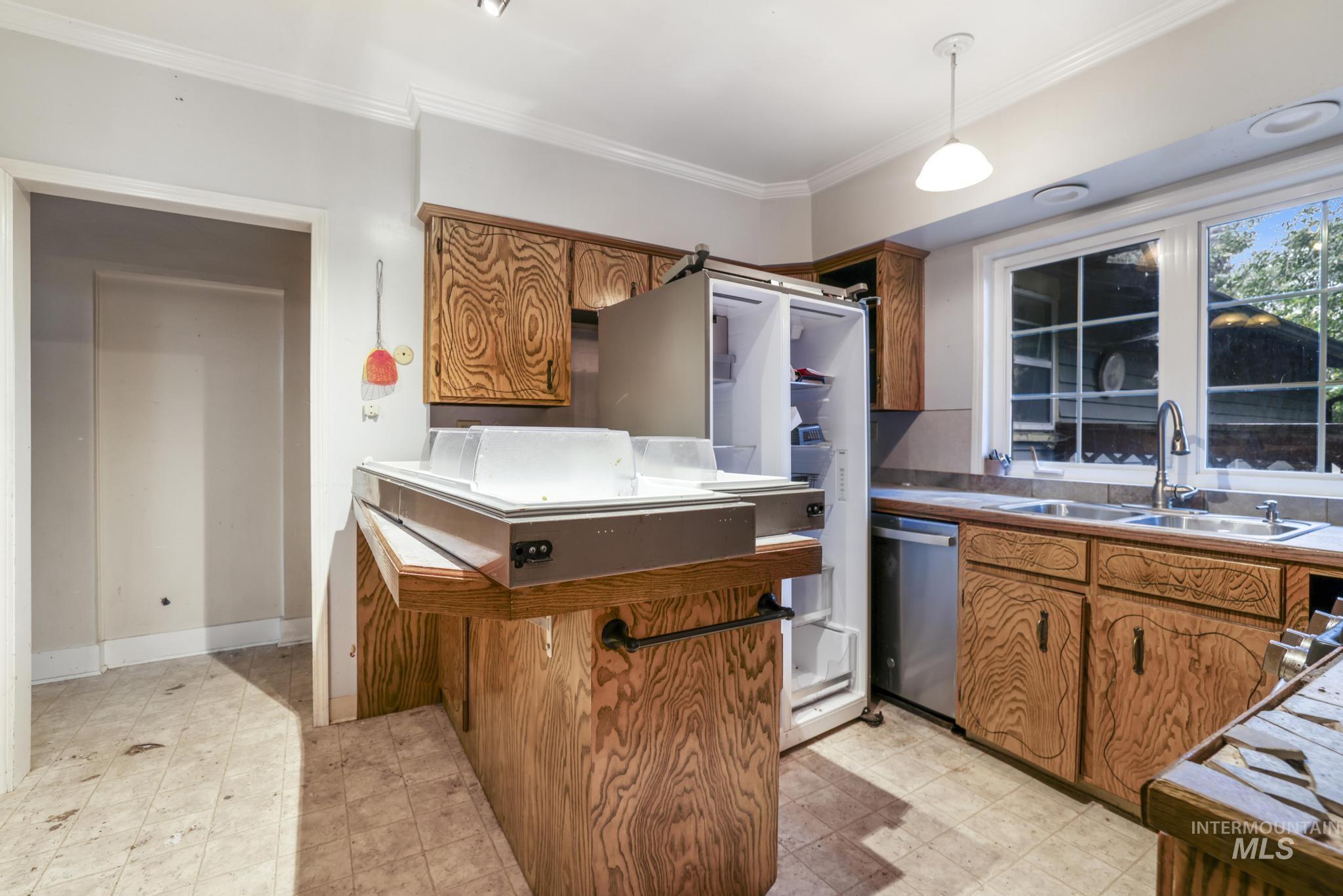 Kitchen featuring brown cabinets, ornamental molding, stainless steel dishwasher, light floors, and decorative light fixtures