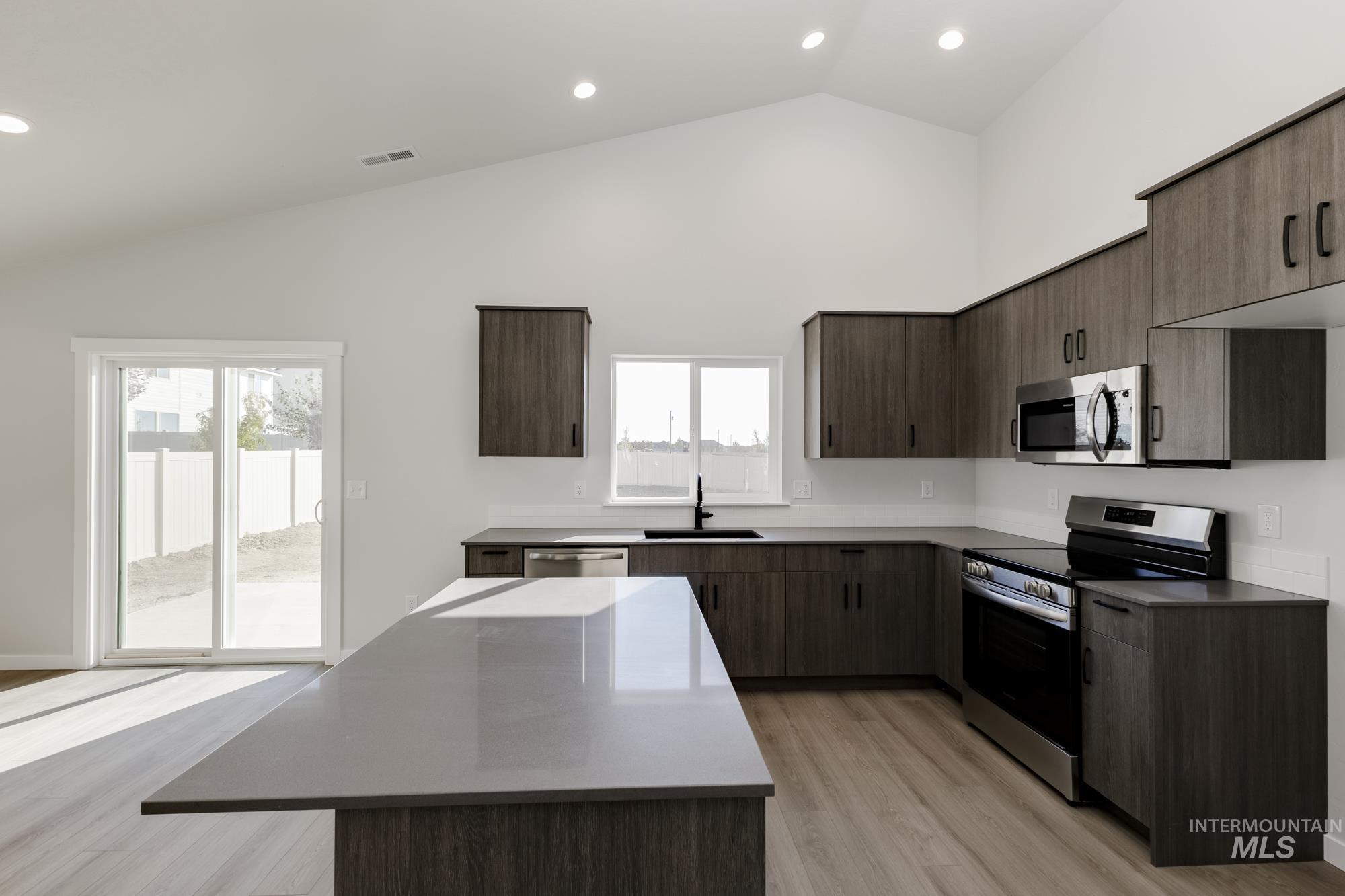 Kitchen with dark brown cabinetry, appliances with stainless steel finishes, light wood-style flooring, recessed lighting, and high vaulted ceiling