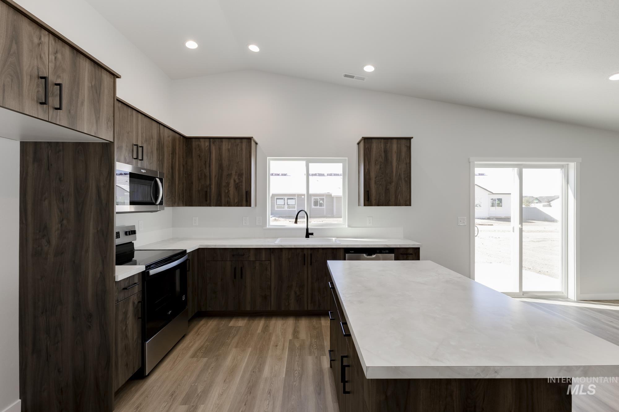 Kitchen with dark brown cabinetry, stainless steel appliances, light countertops, vaulted ceiling, and light wood finished floors