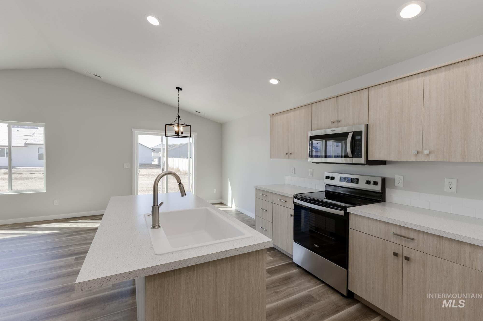 Kitchen featuring light brown cabinetry, appliances with stainless steel finishes, an island with sink, hanging light fixtures, and lofted ceiling