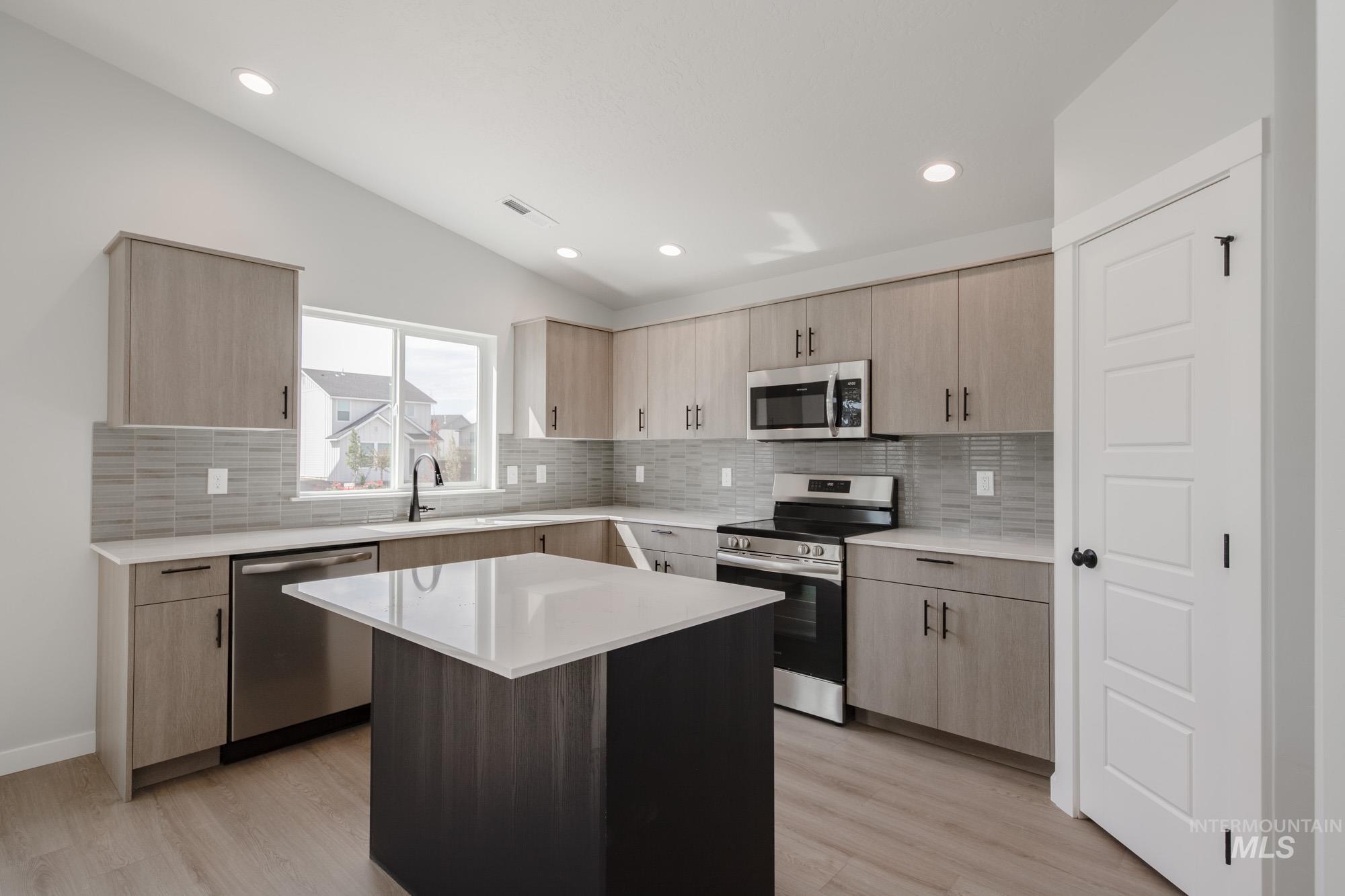 Kitchen with light brown cabinets, appliances with stainless steel finishes, tasteful backsplash, light stone counters, and vaulted ceiling