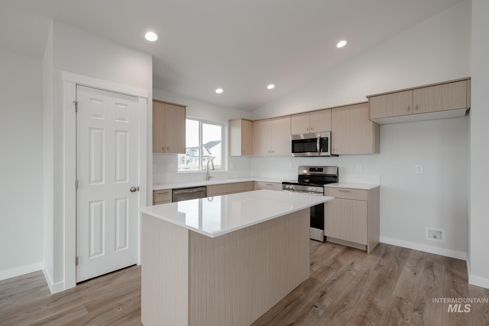 Kitchen featuring light brown cabinetry, appliances with stainless steel finishes, vaulted ceiling, light wood-style floors, and a kitchen island