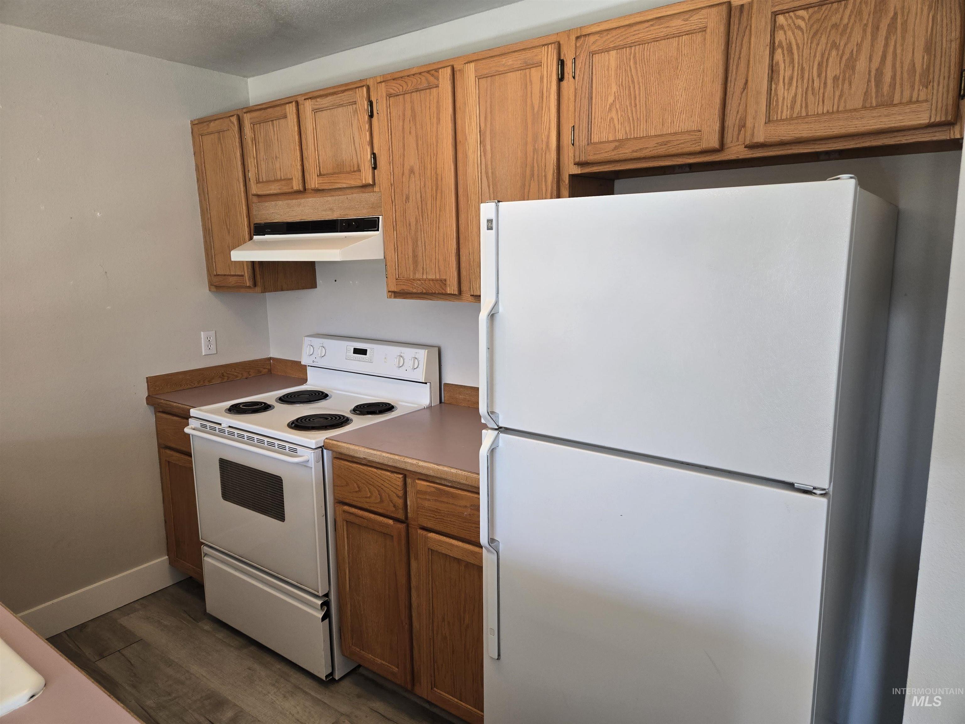 Kitchen featuring white appliances, dark wood finished floors, under cabinet range hood, brown cabinets, and dark countertops