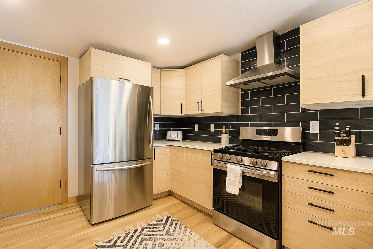 Kitchen with stainless steel appliances, light brown cabinetry, wall chimney range hood, modern cabinets, and tasteful backsplash