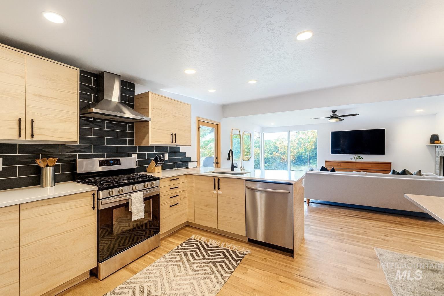Kitchen with light brown cabinets, modern cabinets, appliances with stainless steel finishes, open floor plan, and wall chimney exhaust hood