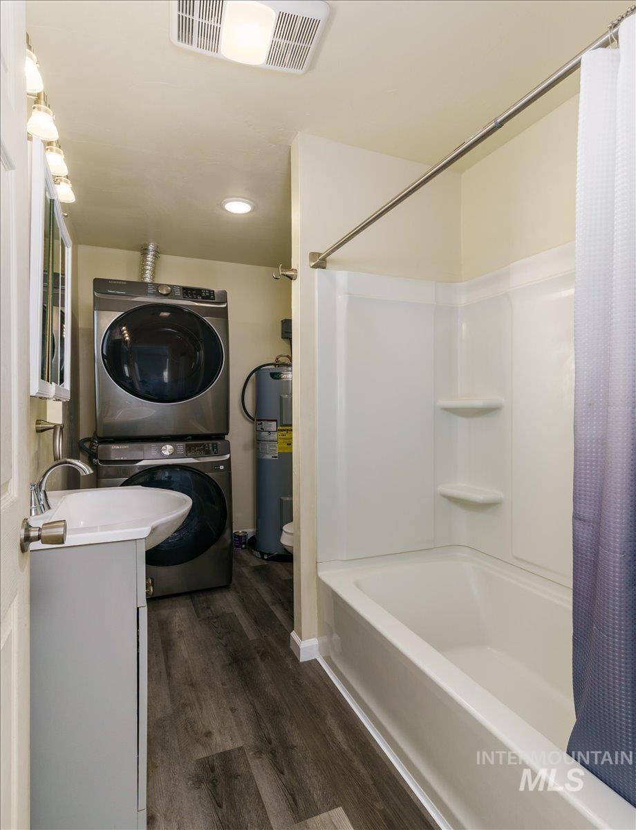 Bathroom featuring vanity, dark wood finished floors, estacked washer and dryer, shower / tub combo with curtain, and water heater