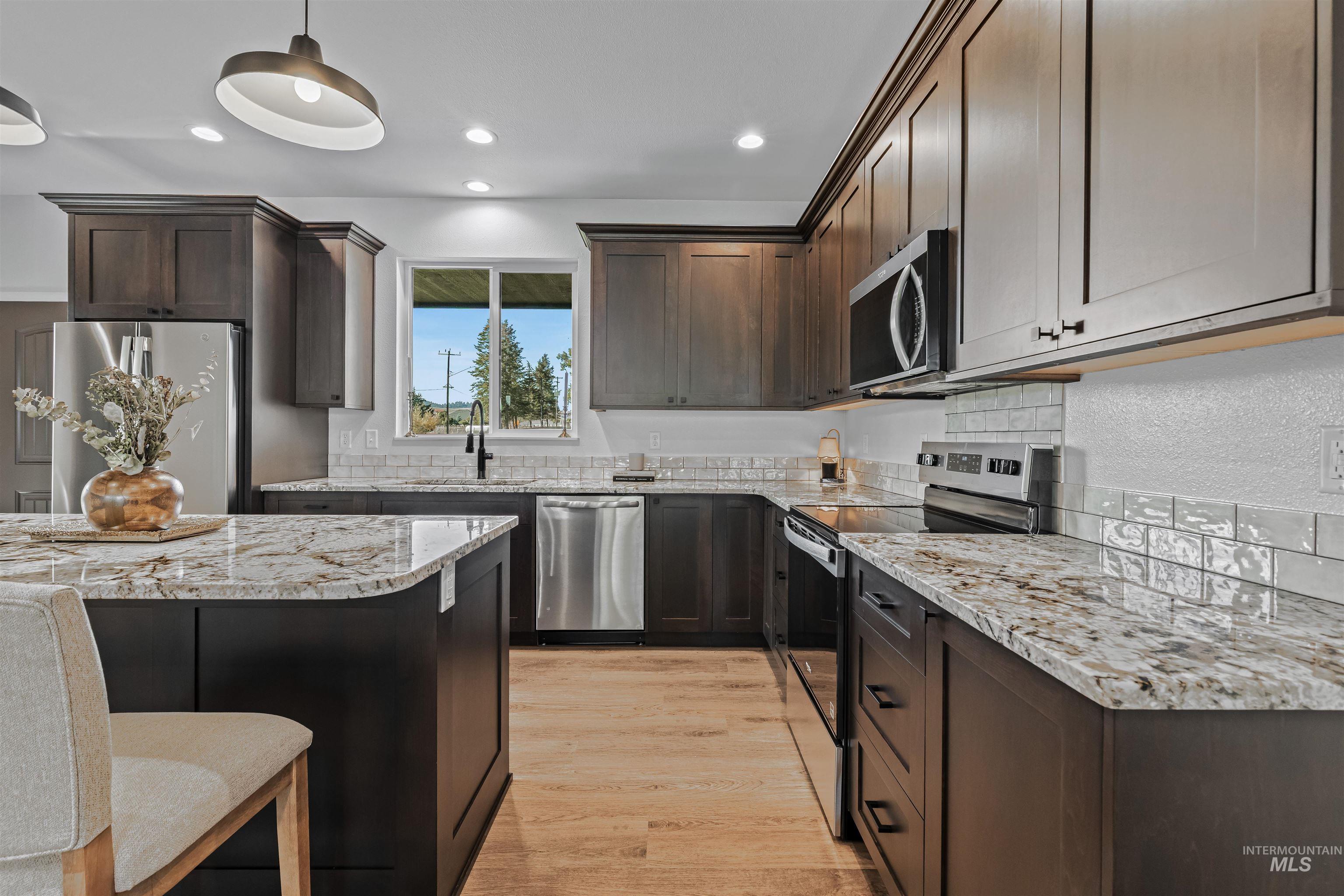 Kitchen featuring appliances with stainless steel finishes, light stone countertops, dark brown cabinetry, hanging light fixtures, and light wood-style flooring