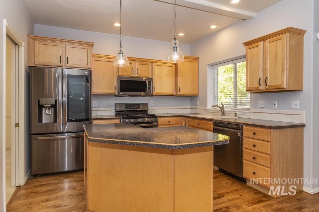 Kitchen featuring stainless steel appliances, hanging light fixtures, dark stone counters, dark wood-type flooring, and light brown cabinetry