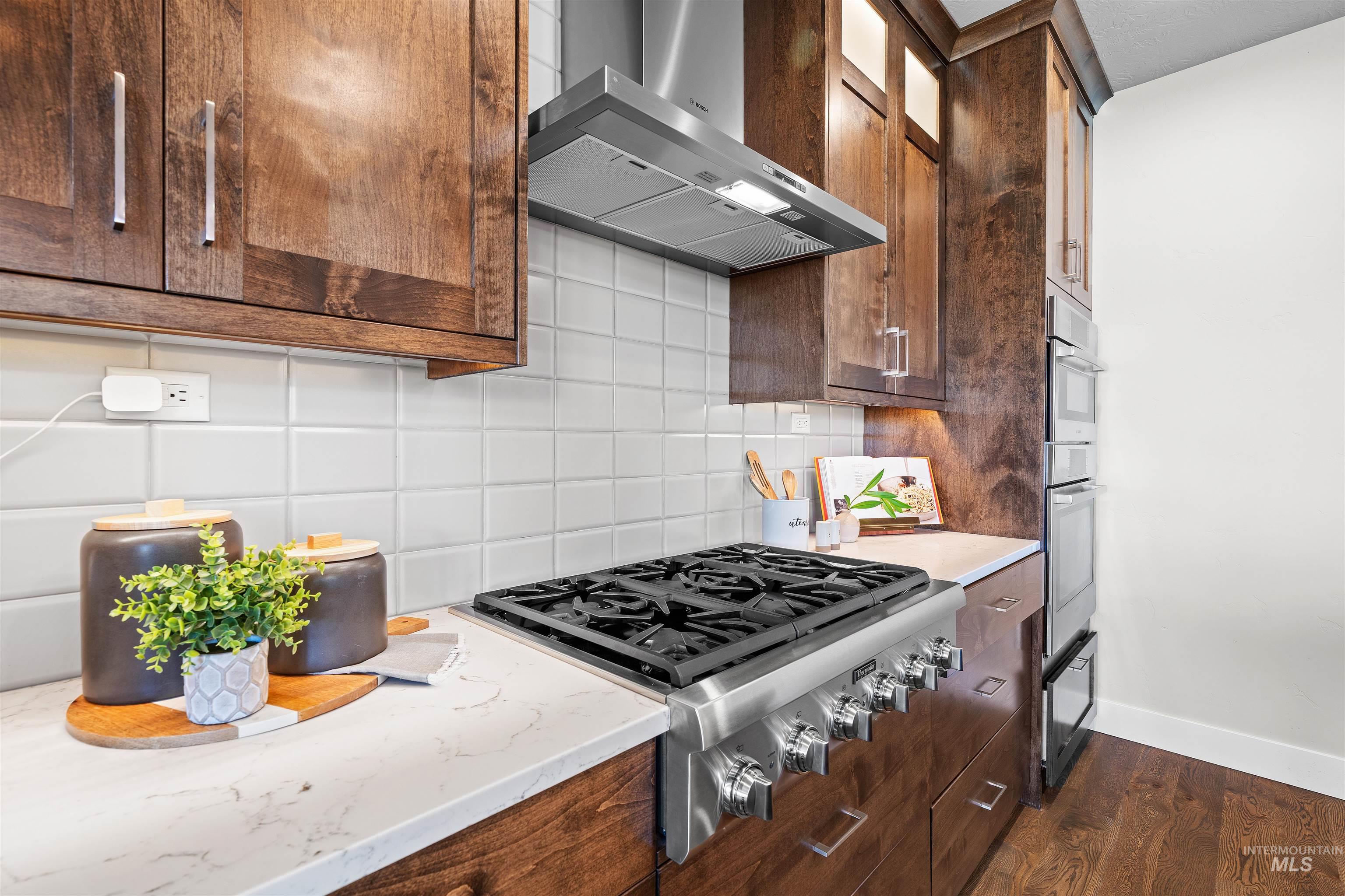 Kitchen featuring wall chimney exhaust hood, tasteful backsplash, appliances with stainless steel finishes, dark wood finished floors, and light stone countertops