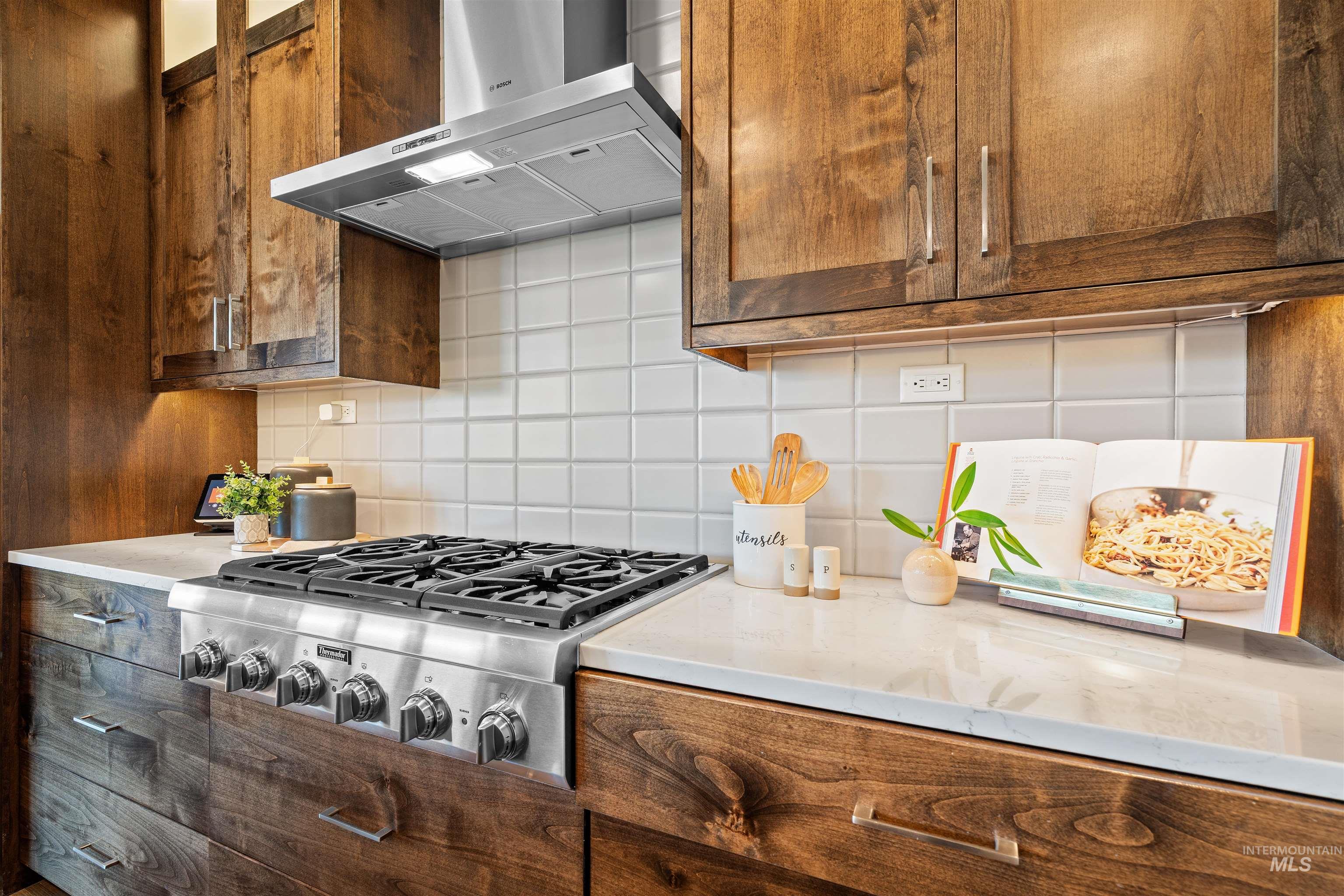 Kitchen with wall chimney range hood, decorative backsplash, stainless steel gas stovetop, light stone countertops, and brown cabinetry