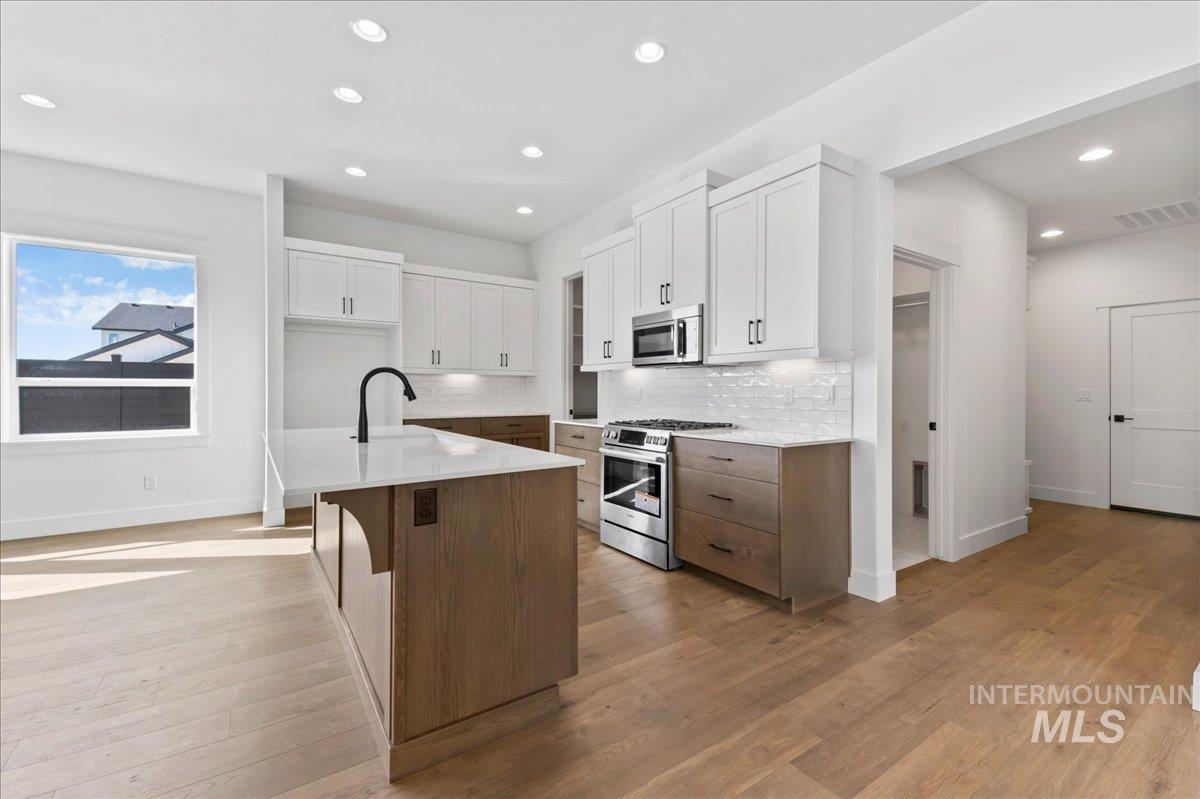 Kitchen with white cabinetry, decorative backsplash, stainless steel appliances, brown cabinets, and a center island with sink