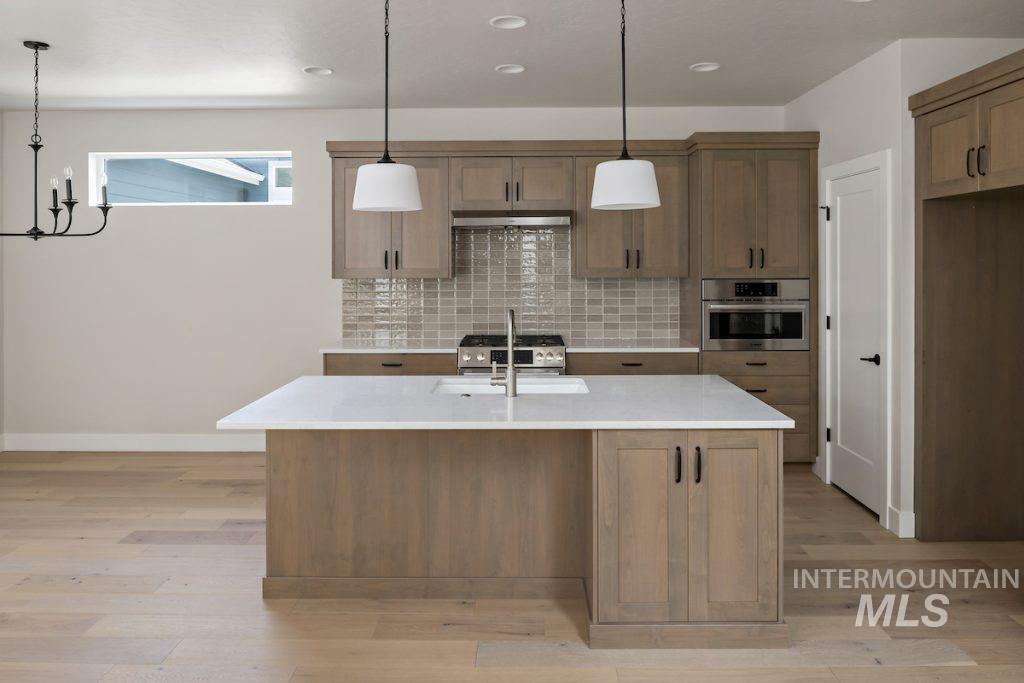 Kitchen with decorative light fixtures, brown cabinetry, an island with sink, light stone counters, and stainless steel appliances