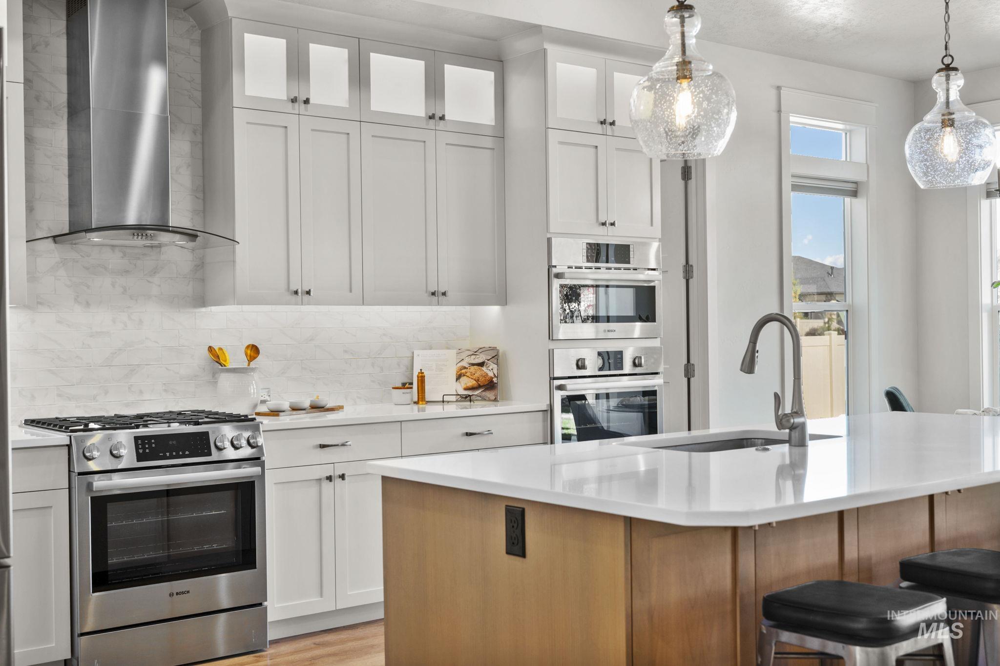 Kitchen featuring stainless steel appliances, wall chimney exhaust hood, decorative light fixtures, decorative backsplash, and white cabinetry