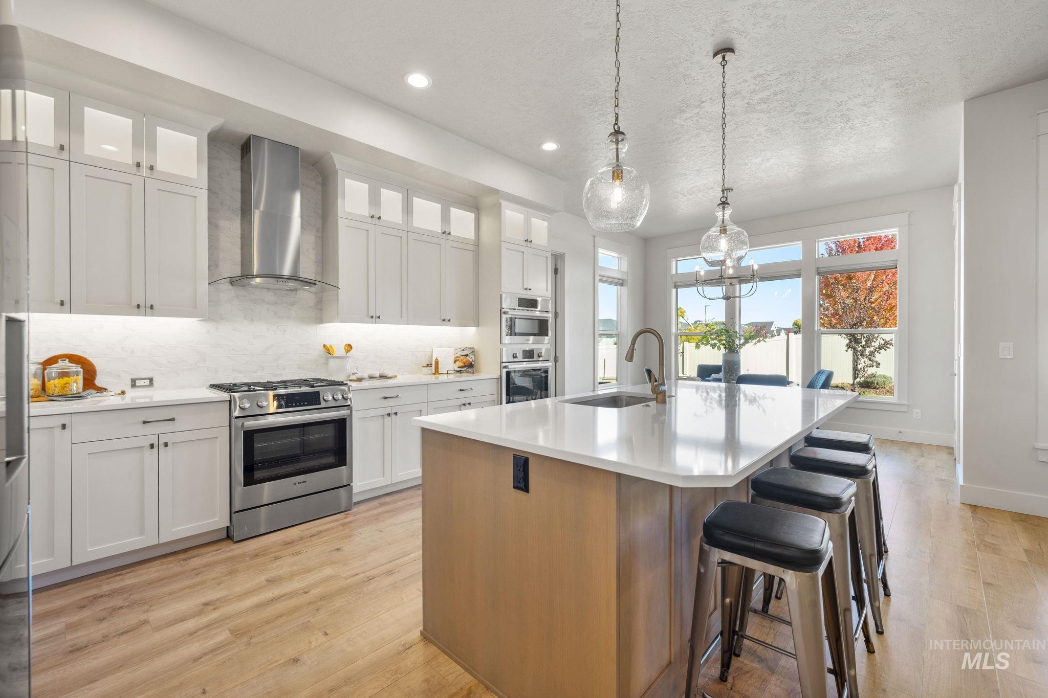 Kitchen with stainless steel appliances, wall chimney exhaust hood, hanging light fixtures, white cabinets, and tasteful backsplash