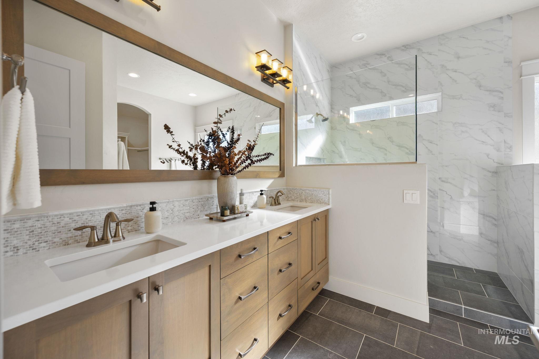 Bathroom featuring a marble finish shower, double vanity, dark tile patterned floors, and backsplash