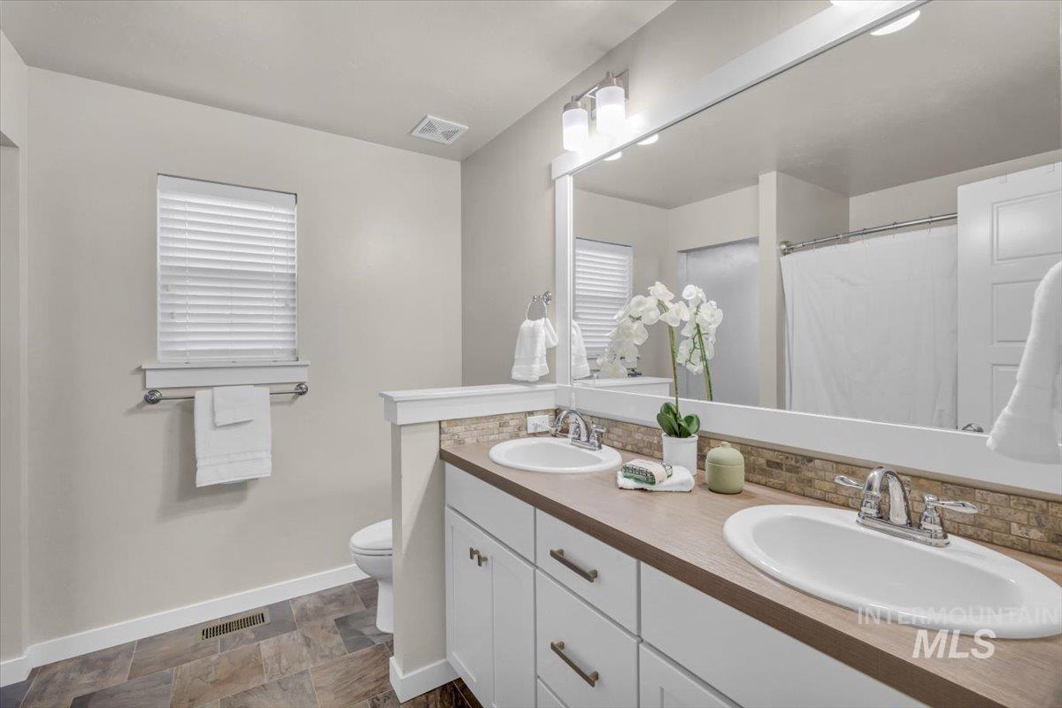 Full bath with double vanity, curtained shower, backsplash, and stone finish floors