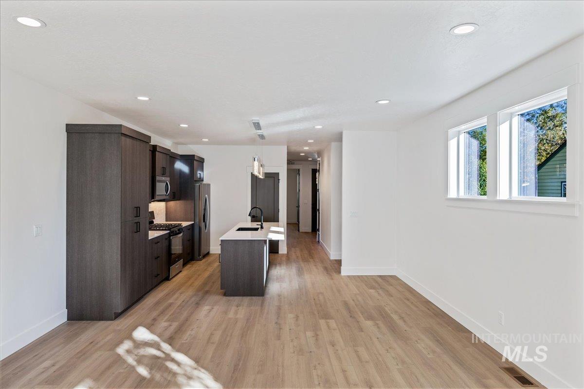 Kitchen featuring an island with sink, dark brown cabinetry, stainless steel appliances, light wood finished floors, and decorative light fixtures