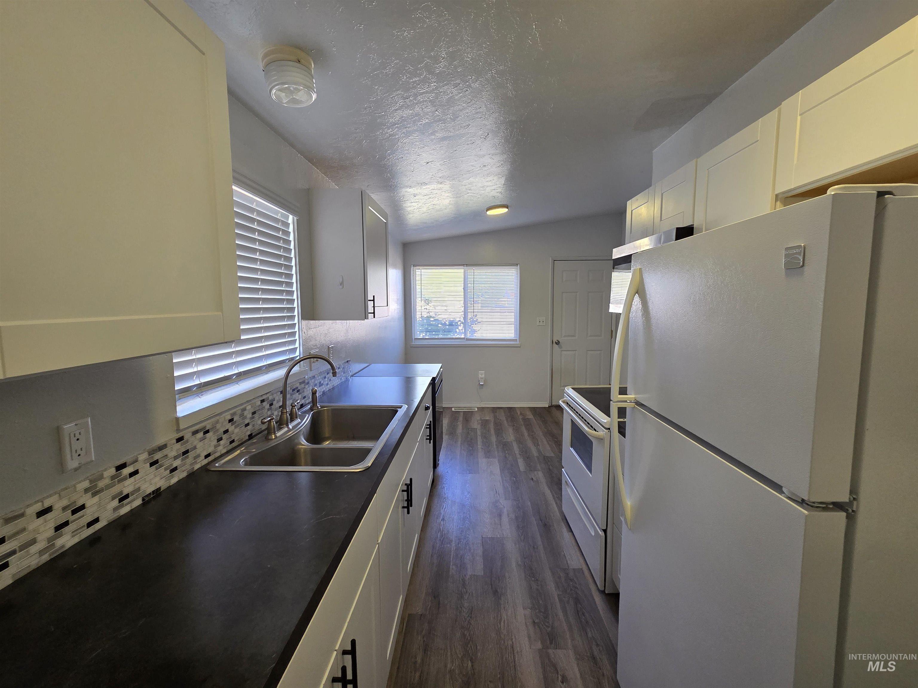 Kitchen with white appliances, backsplash, a textured ceiling, white cabinetry, and vaulted ceiling