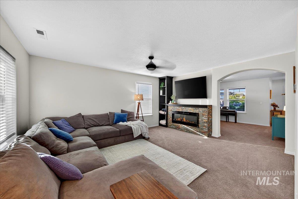 Living area with carpet flooring, a stone fireplace, arched walkways, a textured ceiling, and a ceiling fan