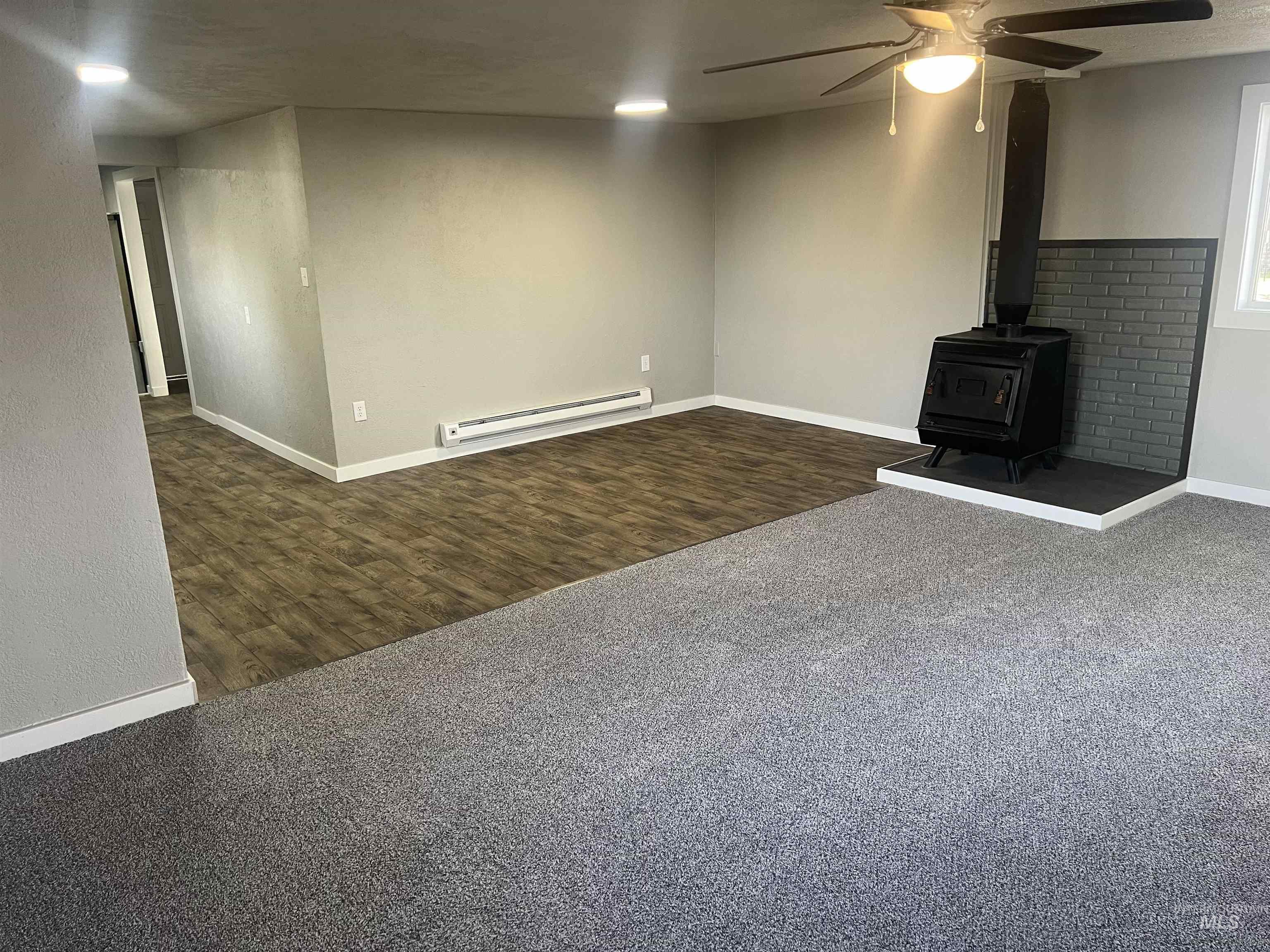 Unfurnished living room featuring a wood stove, dark colored carpet, a ceiling fan, a baseboard radiator, and dark wood-style flooring