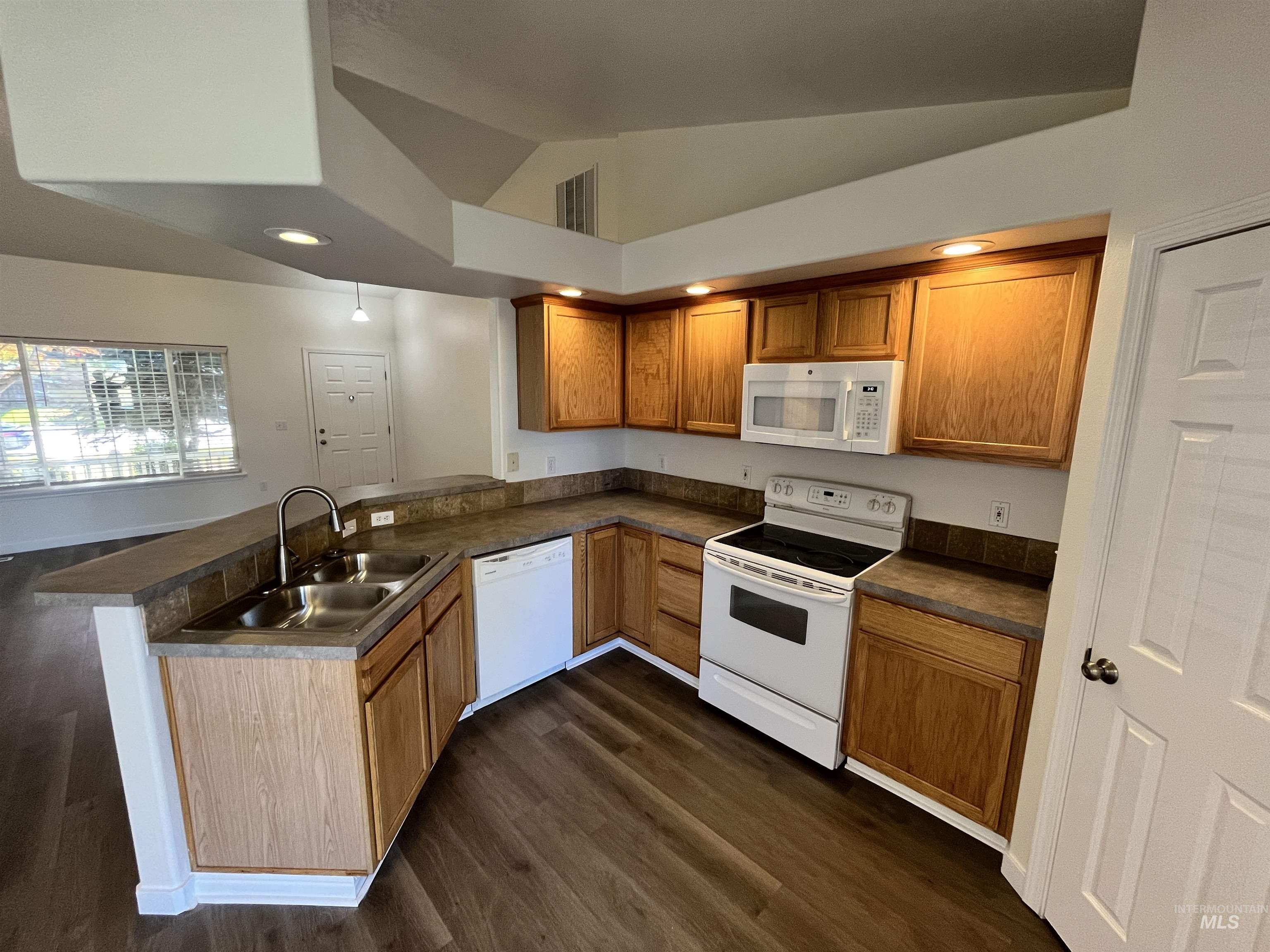 Kitchen featuring a peninsula, white appliances, dark wood finished floors, dark countertops, and brown cabinets