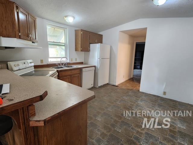 Kitchen featuring brown cabinetry, light countertops, white appliances, under cabinet range hood, and vaulted ceiling