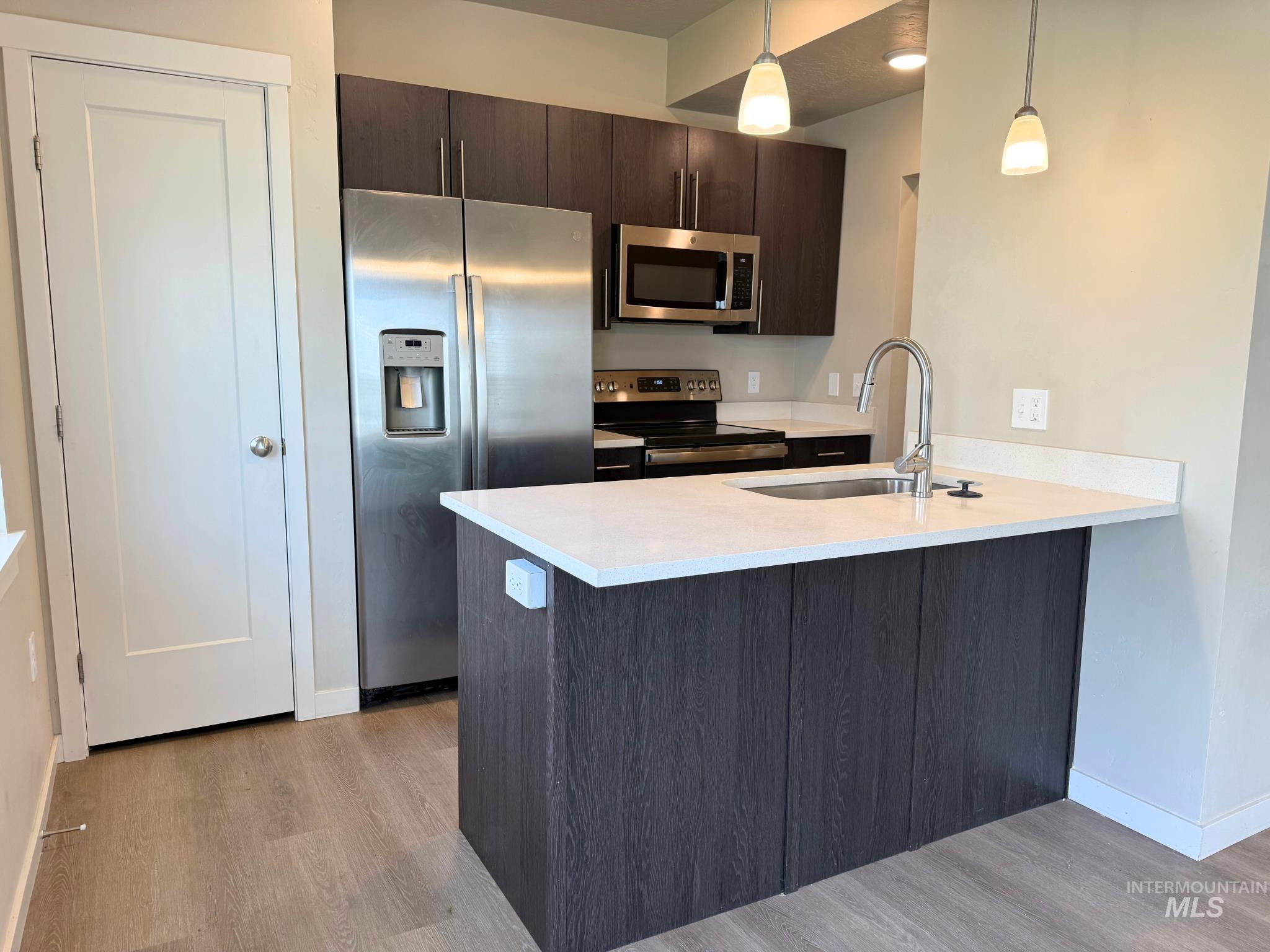 Kitchen with appliances with stainless steel finishes, dark brown cabinets, a peninsula, hanging light fixtures, and light wood finished floors
