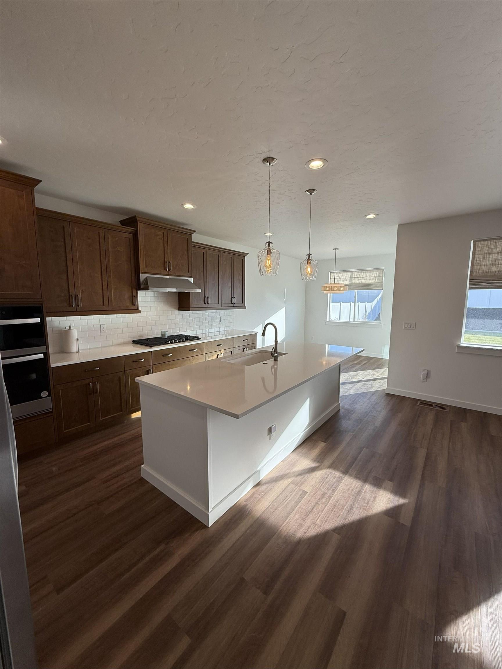 Kitchen with an island with sink, backsplash, pendant lighting, dark wood-type flooring, and double oven