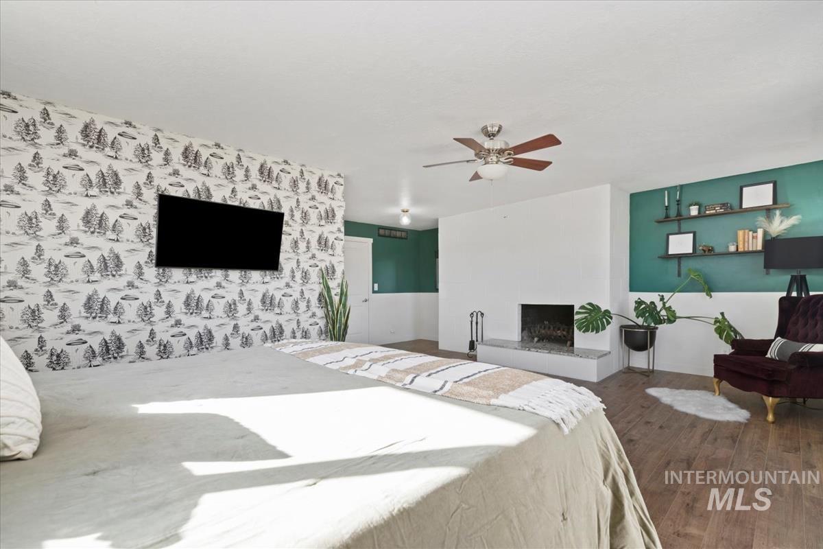 Bedroom featuring wood finished floors, a fireplace with raised hearth, ceiling fan, and wallpapered walls