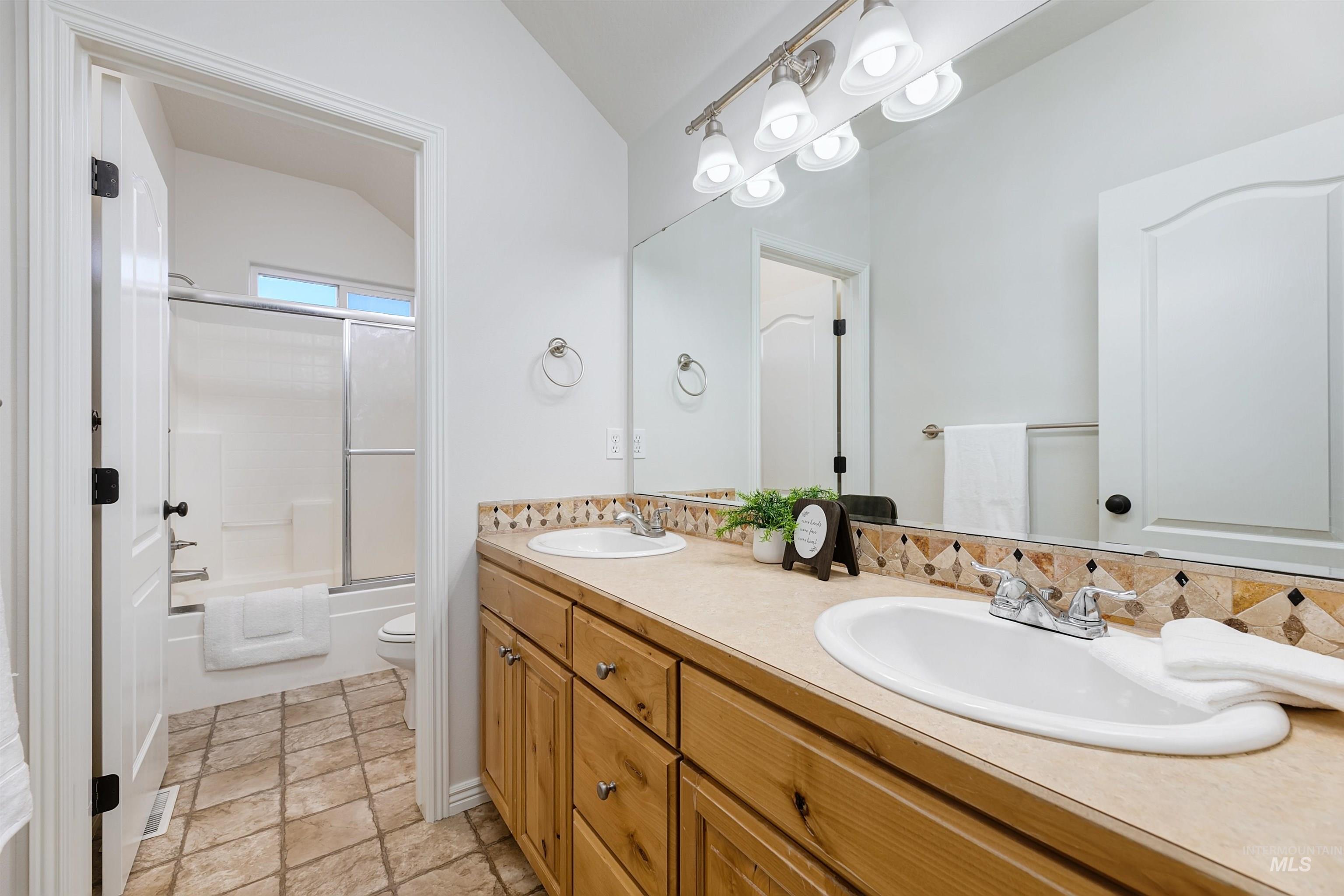 Bathroom with double vanity, light stone finish floors, bath / shower combo with glass door, and vaulted ceiling