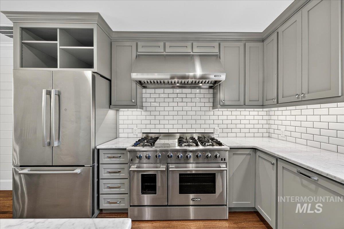 Kitchen featuring gray cabinetry, stainless steel appliances, wall chimney range hood, light stone counters, and open shelves