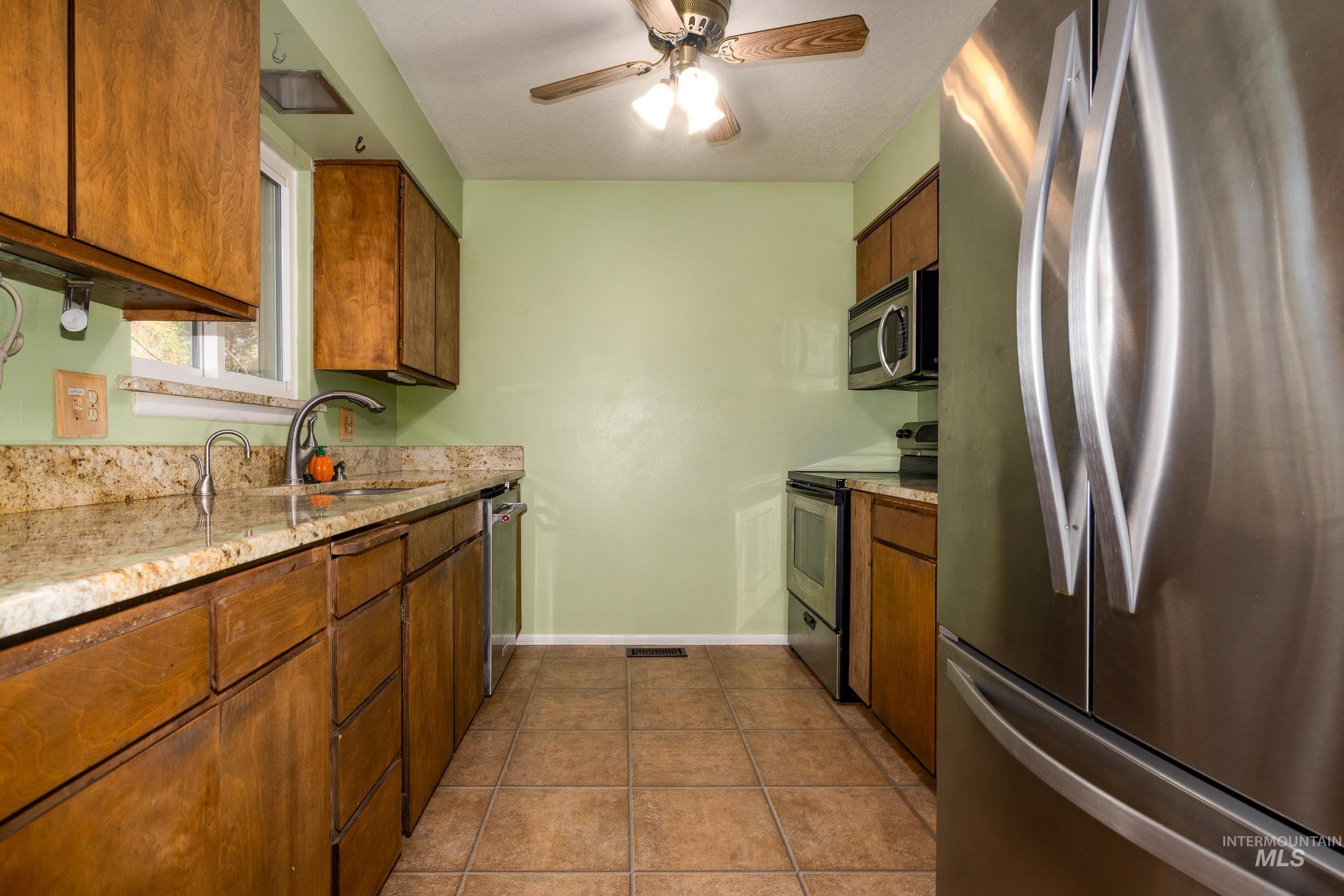 Kitchen featuring stainless steel appliances, brown cabinetry, dark tile patterned flooring, ceiling fan, and light stone counters