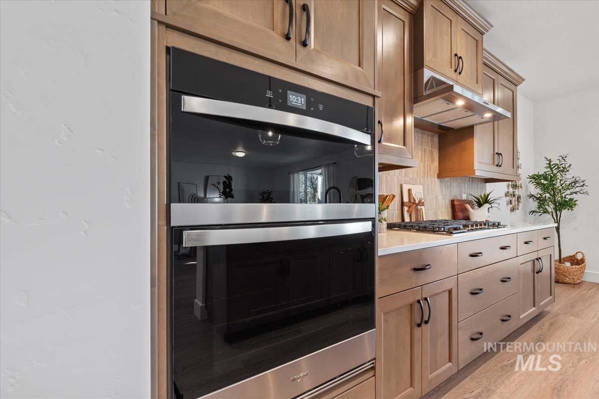 Kitchen with stainless steel appliances, light wood-style flooring, under cabinet range hood, backsplash, and light stone countertops