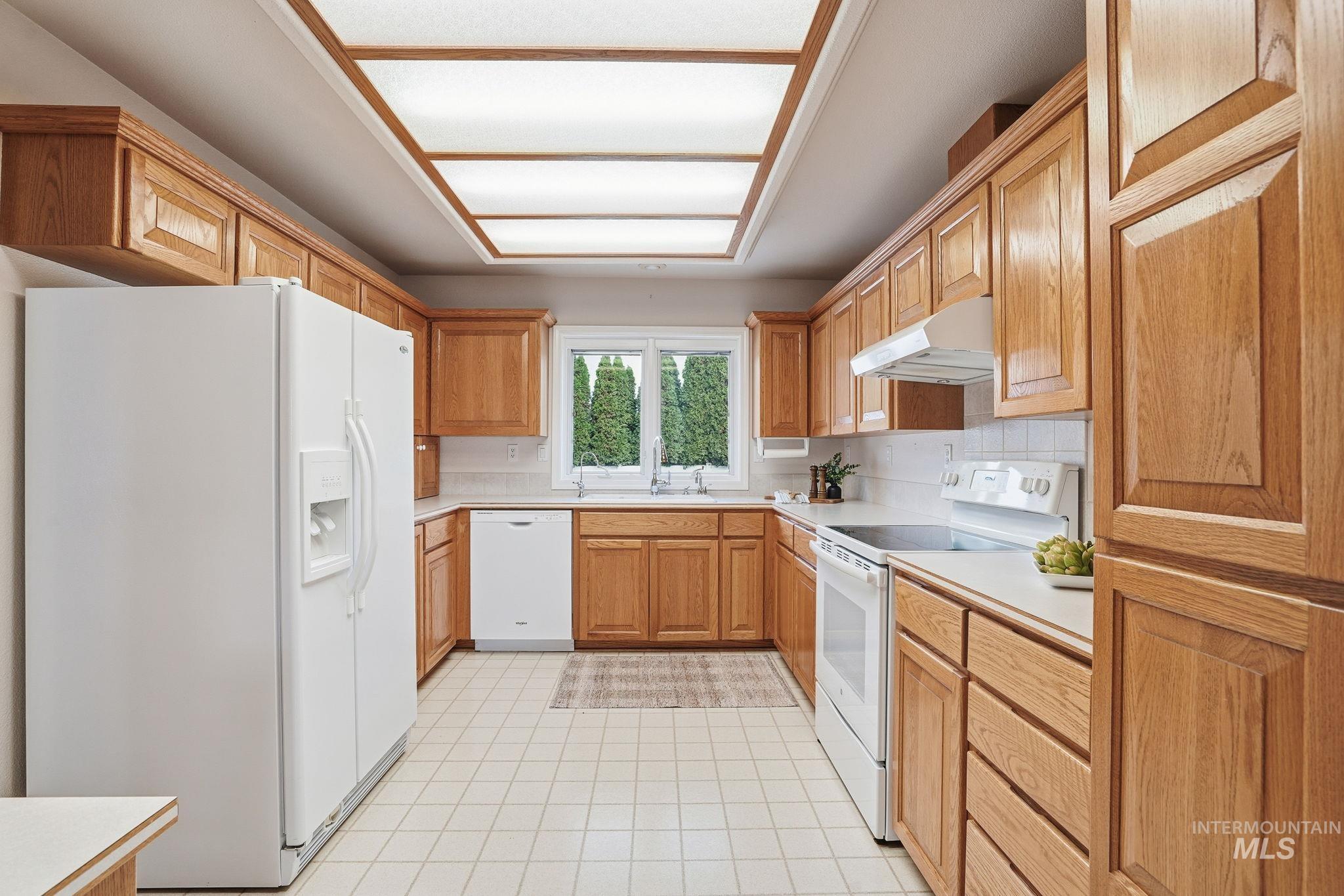 Kitchen featuring white appliances, light countertops, under cabinet range hood, decorative backsplash, and light tile patterned floors