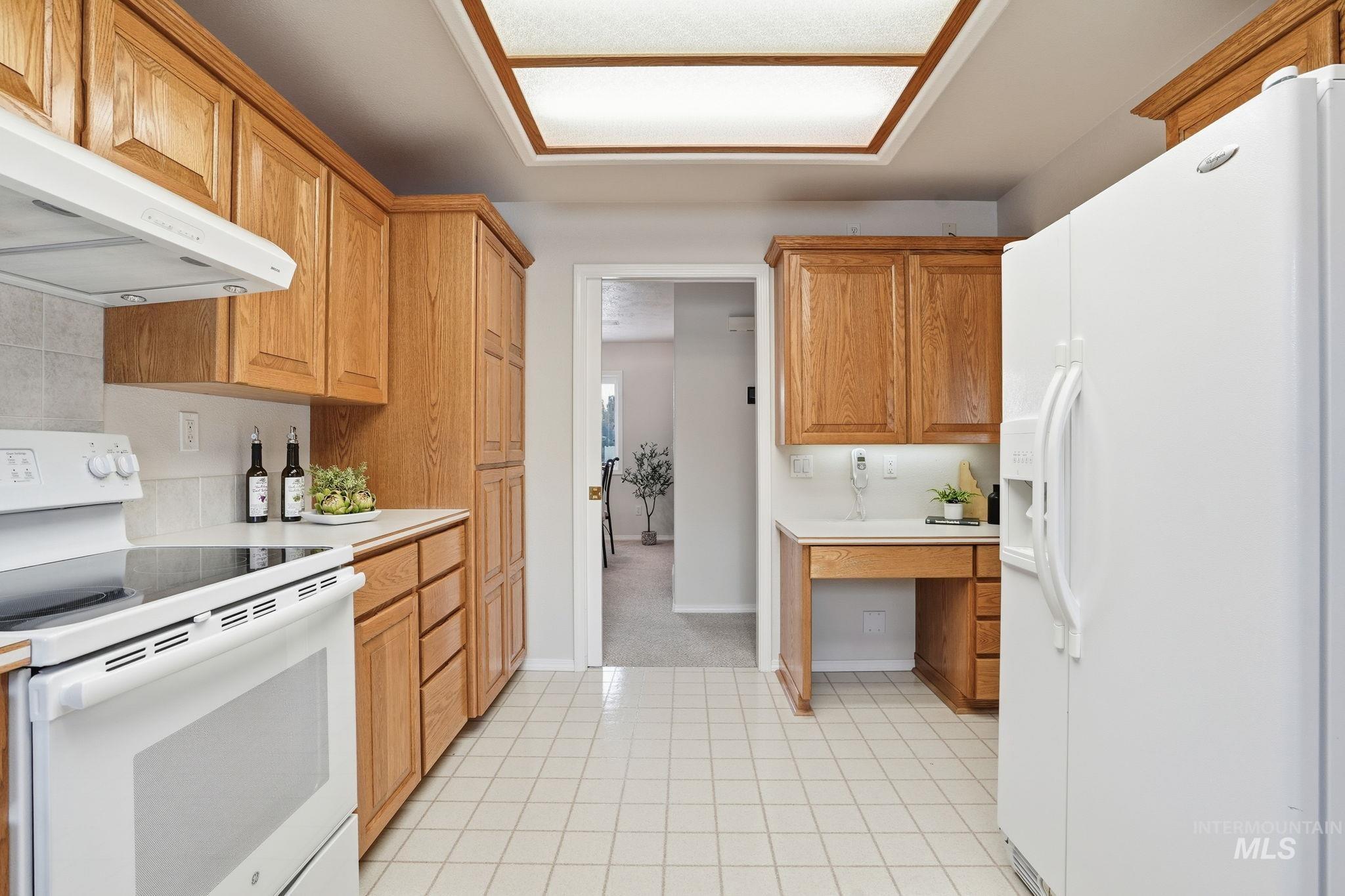 Kitchen with white appliances, light countertops, under cabinet range hood, light tile patterned floors, and brown cabinetry