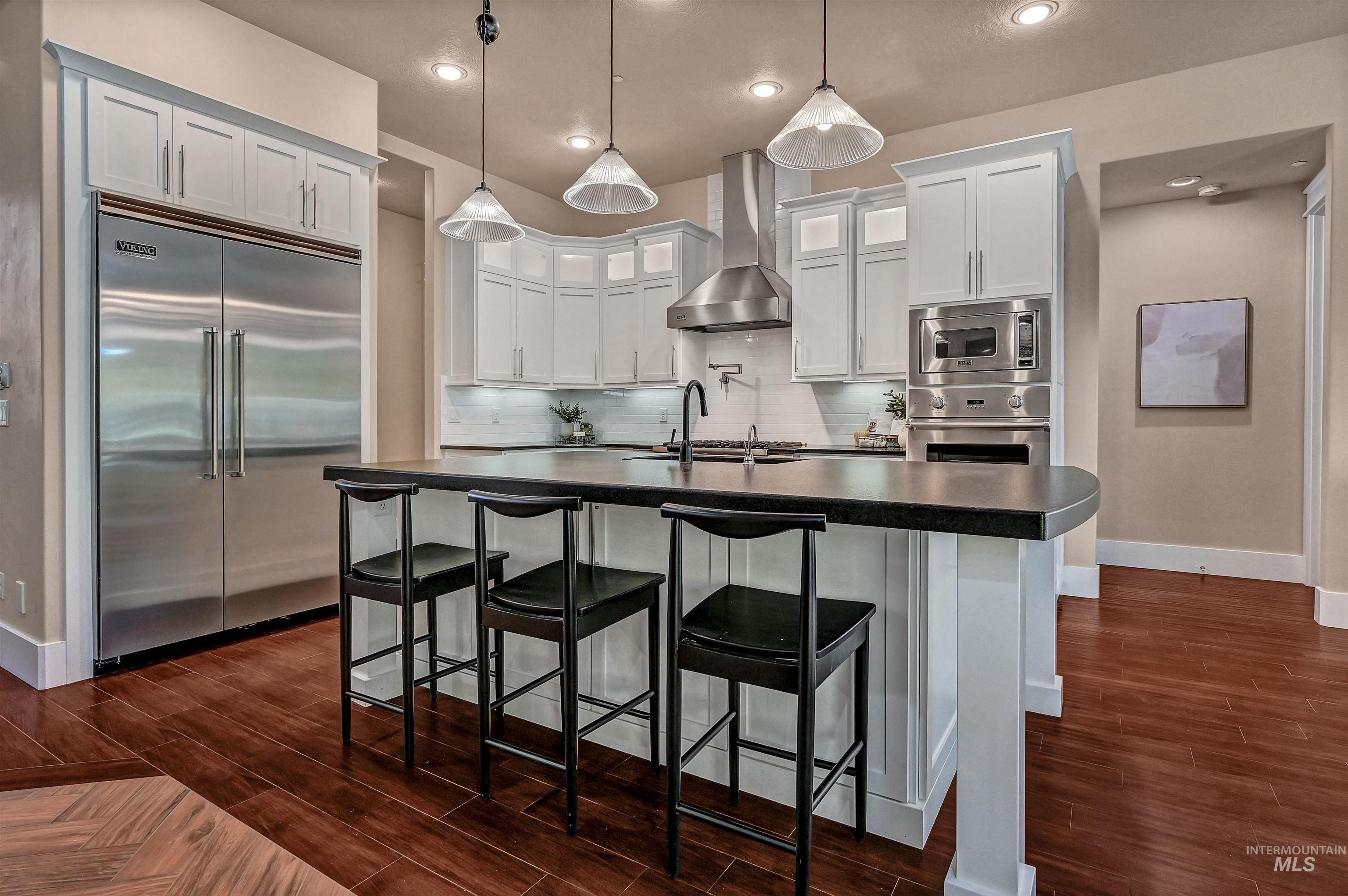 Kitchen with dark countertops, built in appliances, white cabinets, backsplash, and wall chimney range hood