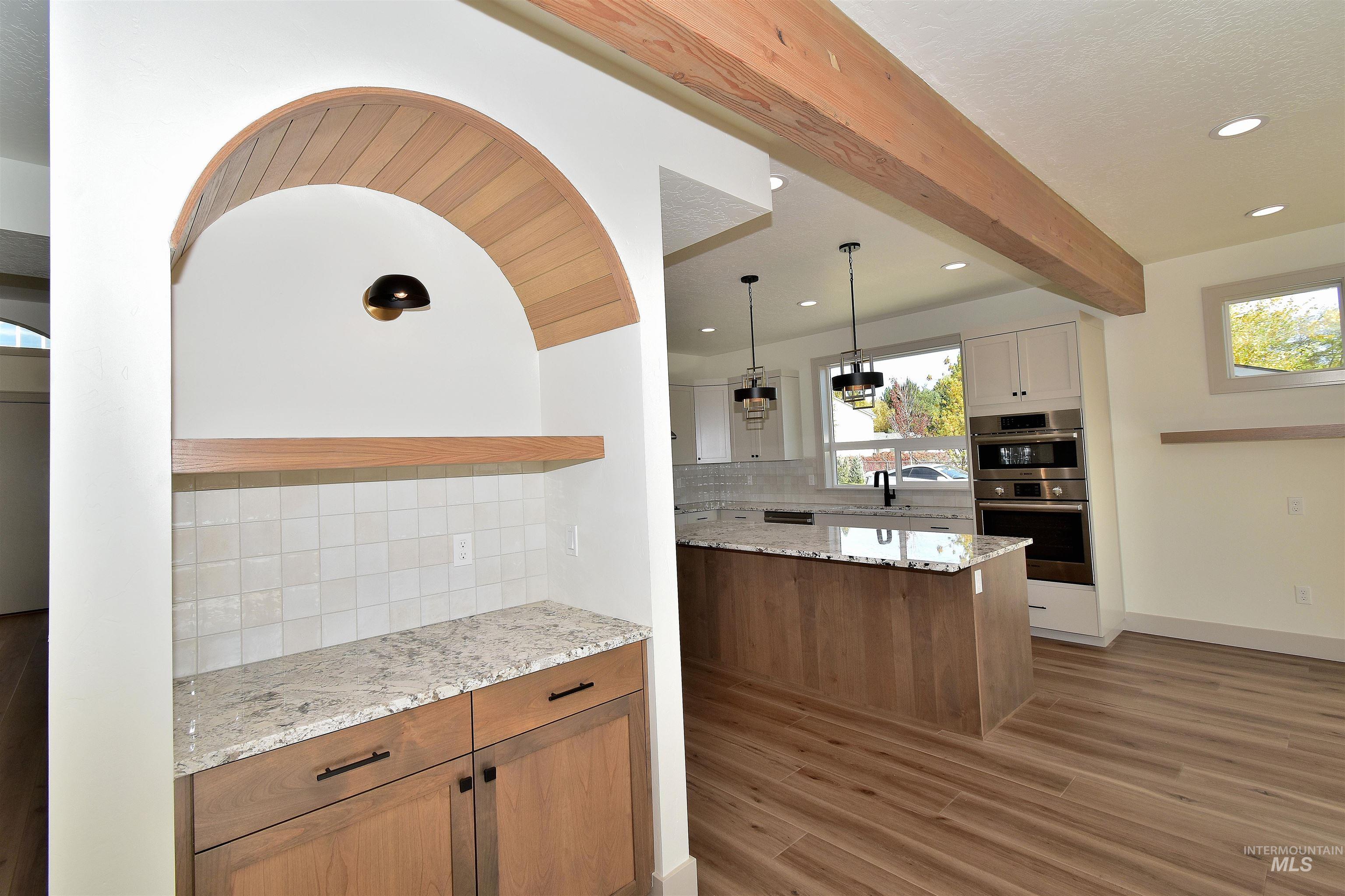 Kitchen featuring beamed ceiling, backsplash, light stone countertops, brown cabinetry, and pendant lighting