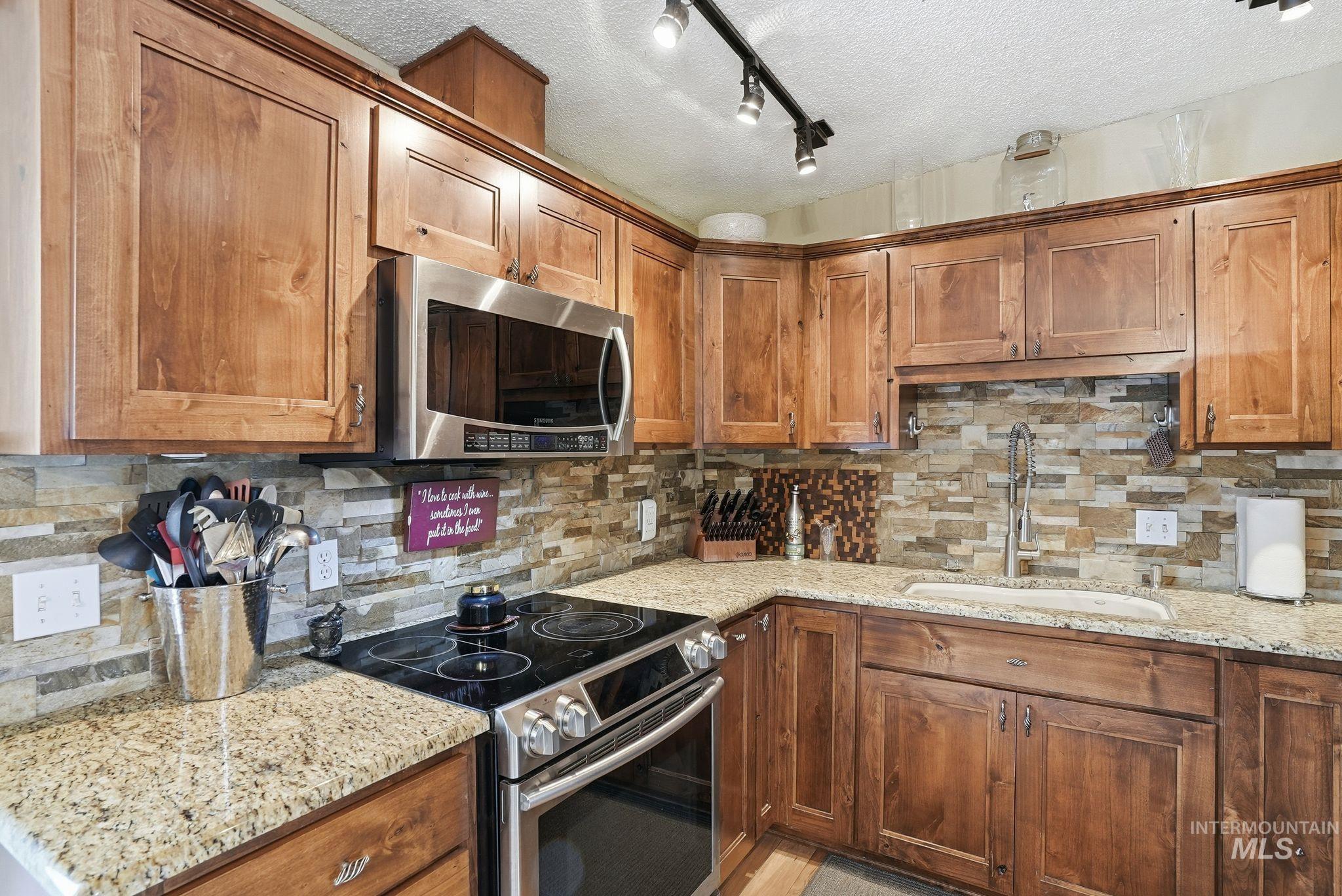 Kitchen featuring stainless steel appliances, decorative backsplash, a textured ceiling, brown cabinetry, and light stone countertops