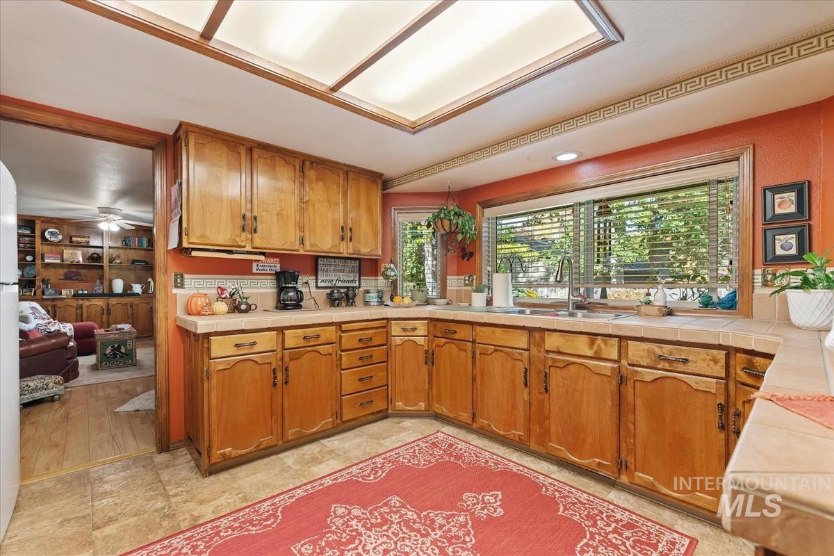 Kitchen with brown cabinetry, tile counters, ceiling fan, and freestanding refrigerator