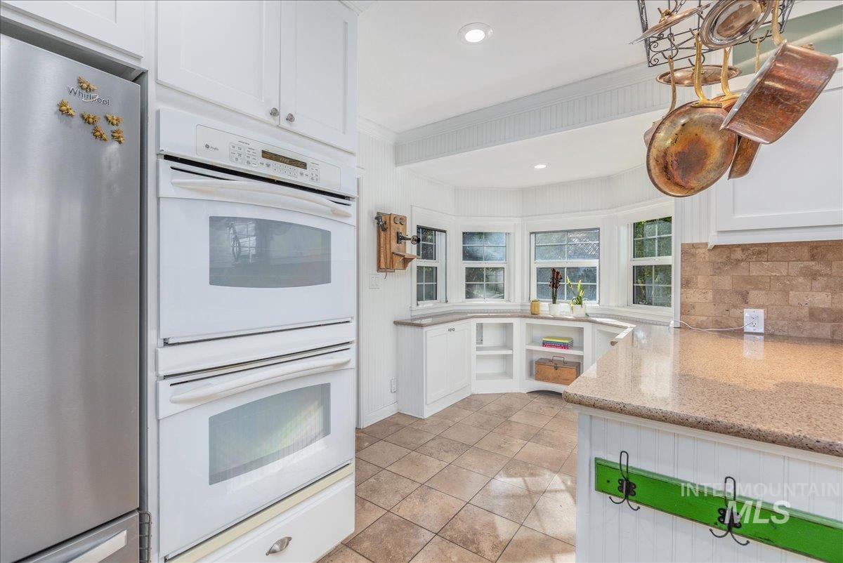Kitchen featuring double oven, white cabinetry, freestanding refrigerator, open shelves, and light stone countertops