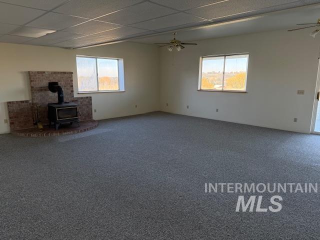 Unfurnished living room with a wood stove, healthy amount of natural light, a drop ceiling, carpet flooring, and ceiling fan