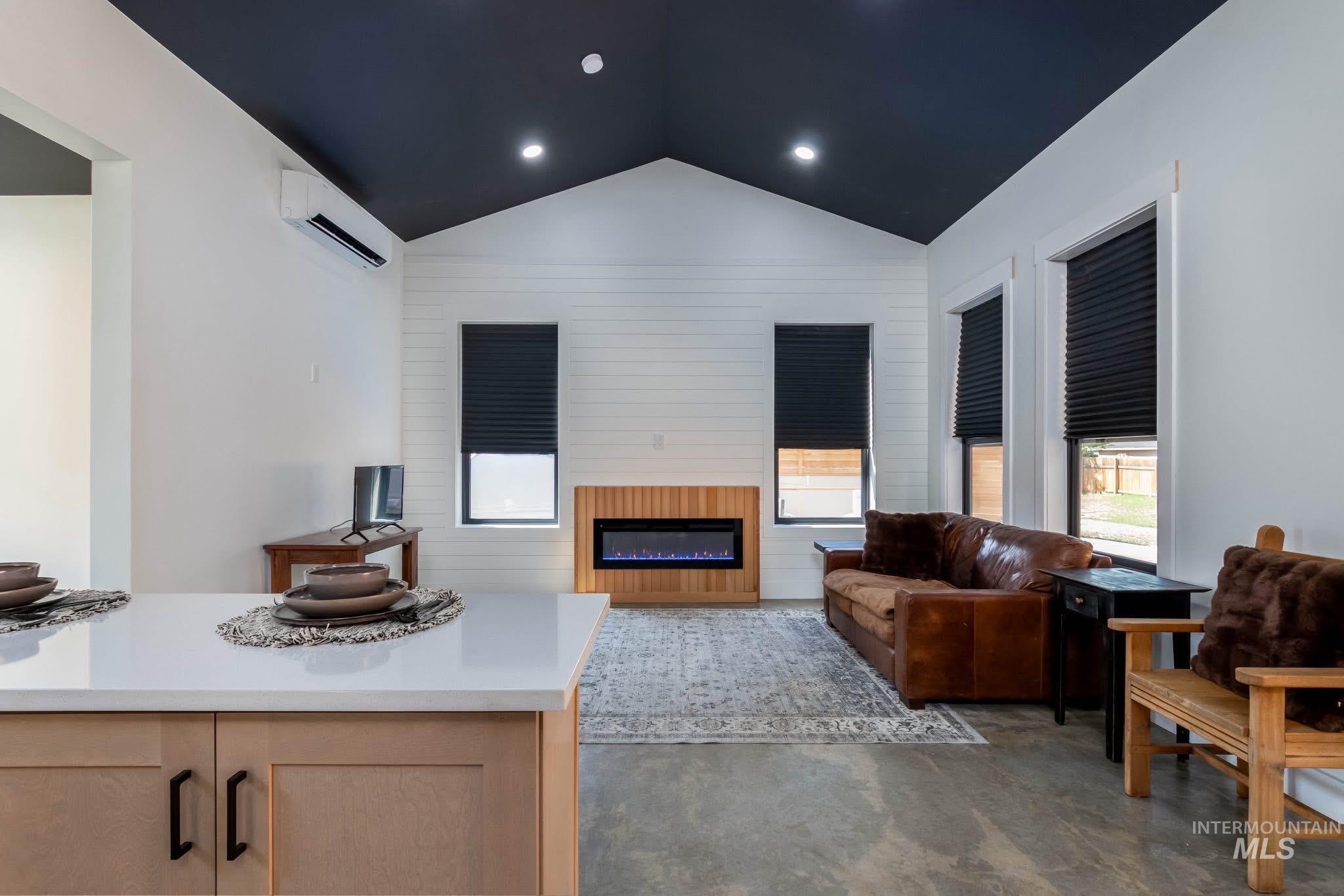 Living area with vaulted ceiling, a glass covered fireplace, wooden walls, concrete floors, and a wall mounted AC