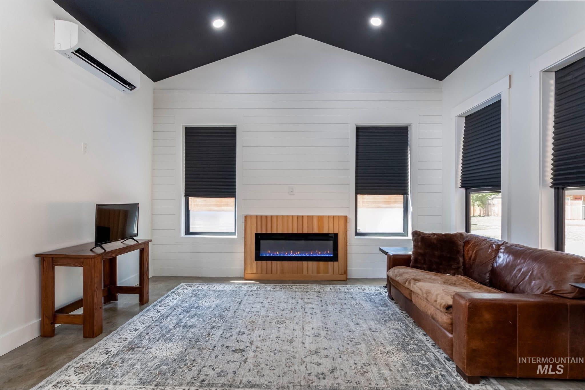 Living area featuring lofted ceiling, a wall mounted AC, a glass covered fireplace, concrete floors, and wooden walls