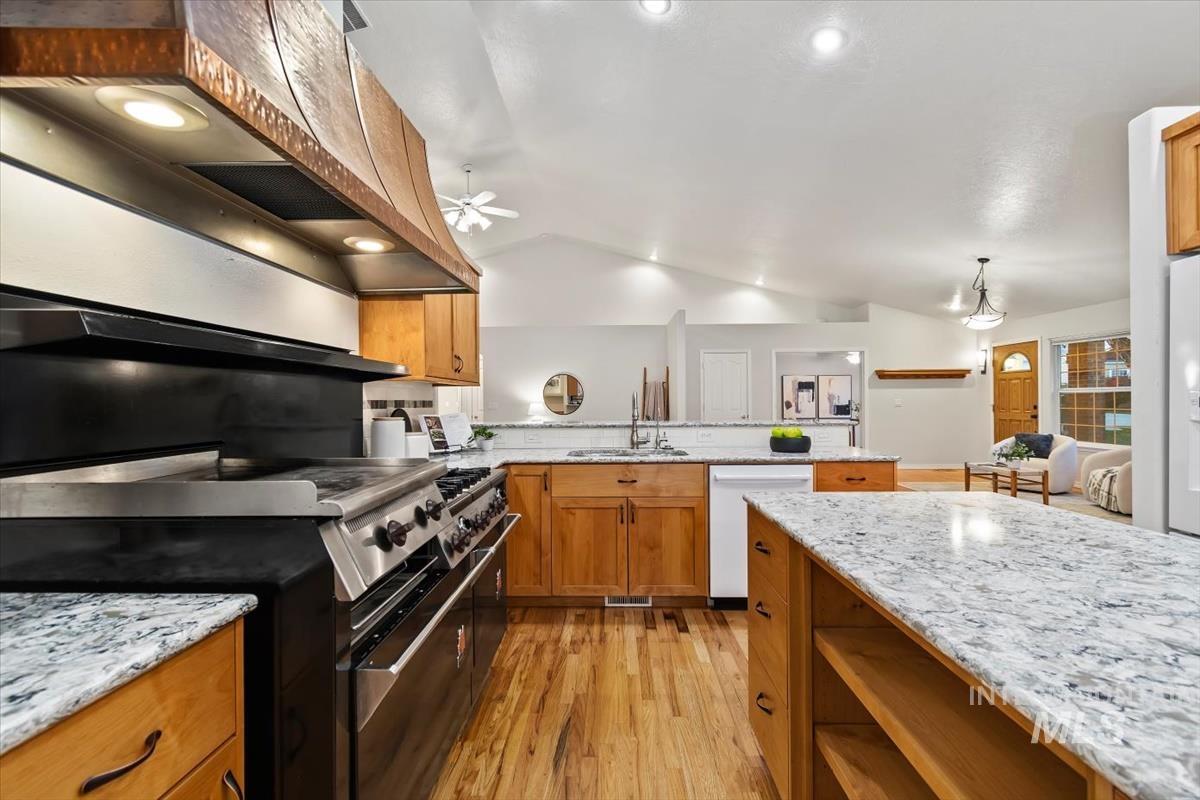 Kitchen with custom exhaust hood, vaulted ceiling, double oven range, brown cabinetry, and light stone countertops