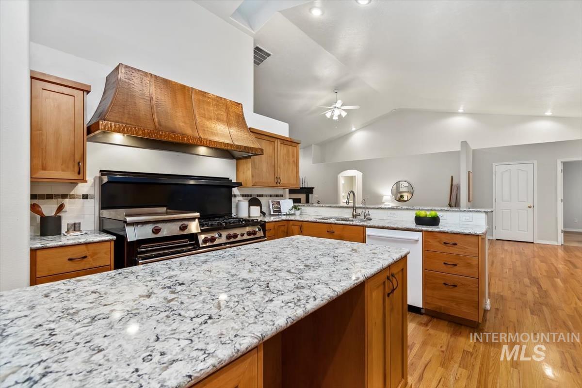 Kitchen featuring brown cabinetry, light stone counters, vaulted ceiling, light wood-style floors, and custom exhaust hood