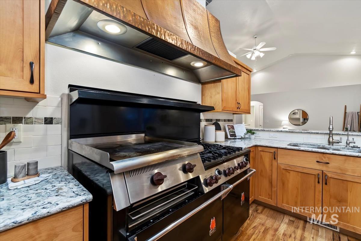 Kitchen featuring under cabinet range hood, decorative backsplash, brown cabinets, light stone counters, and lofted ceiling